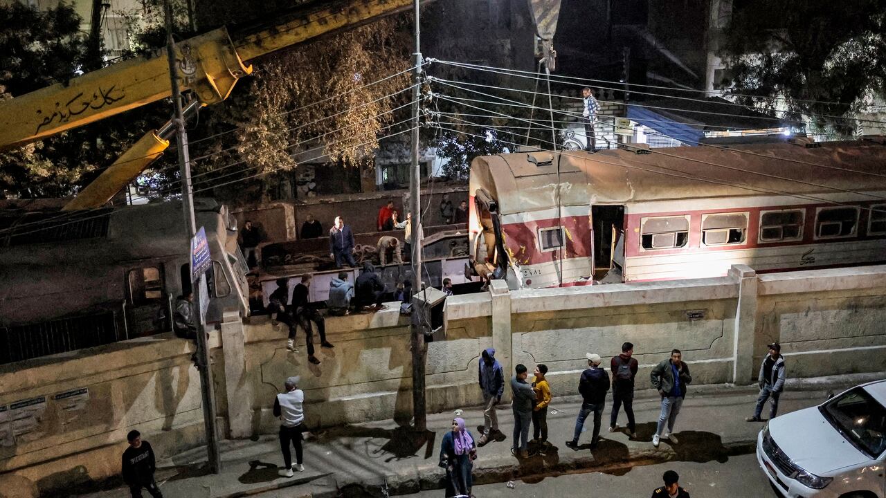 La gente se reúne junto a un muro que cerca la vía del tren mientras se despliega una grúa para levantar un tren descarrilado en la escena de un accidente ferroviario en la ciudad de Qalyub en la provincia de Qalyub, en la región del delta del Nilo de Egipto, al norte de la capital.