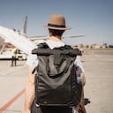 Un joven blanco con sombrero y maleta caminando por el aeropuerto cerca de la pista de despegue
