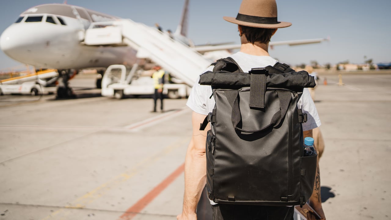 Un joven blanco con sombrero y maleta caminando por el aeropuerto cerca de la pista de despegue