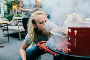 A young man is heating up the barbecue at a party by blowing on the coals.