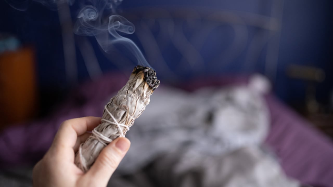 This is a photograph of a burning bundle of sage as a woman’s hand smudges indoors during the shelter in place order during the coronavirus pandemic in Miami, Florida USA.