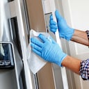 A close up of a Hispanic man wearing disposable gloves as he cleans a stainless steel refrigerator door handle with a disinfectant wipe.