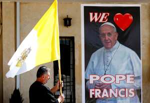 Un sacerdote cristiano sostiene una bandera del vaticano mientras camina ante un cartel del papa Francisco, durante los preparativos para la visita del papa en Mar Youssif Church en Bagdad, Irak, el viernes 26 de febrero de 2021. (AP/Foto/Hadi Mizban)