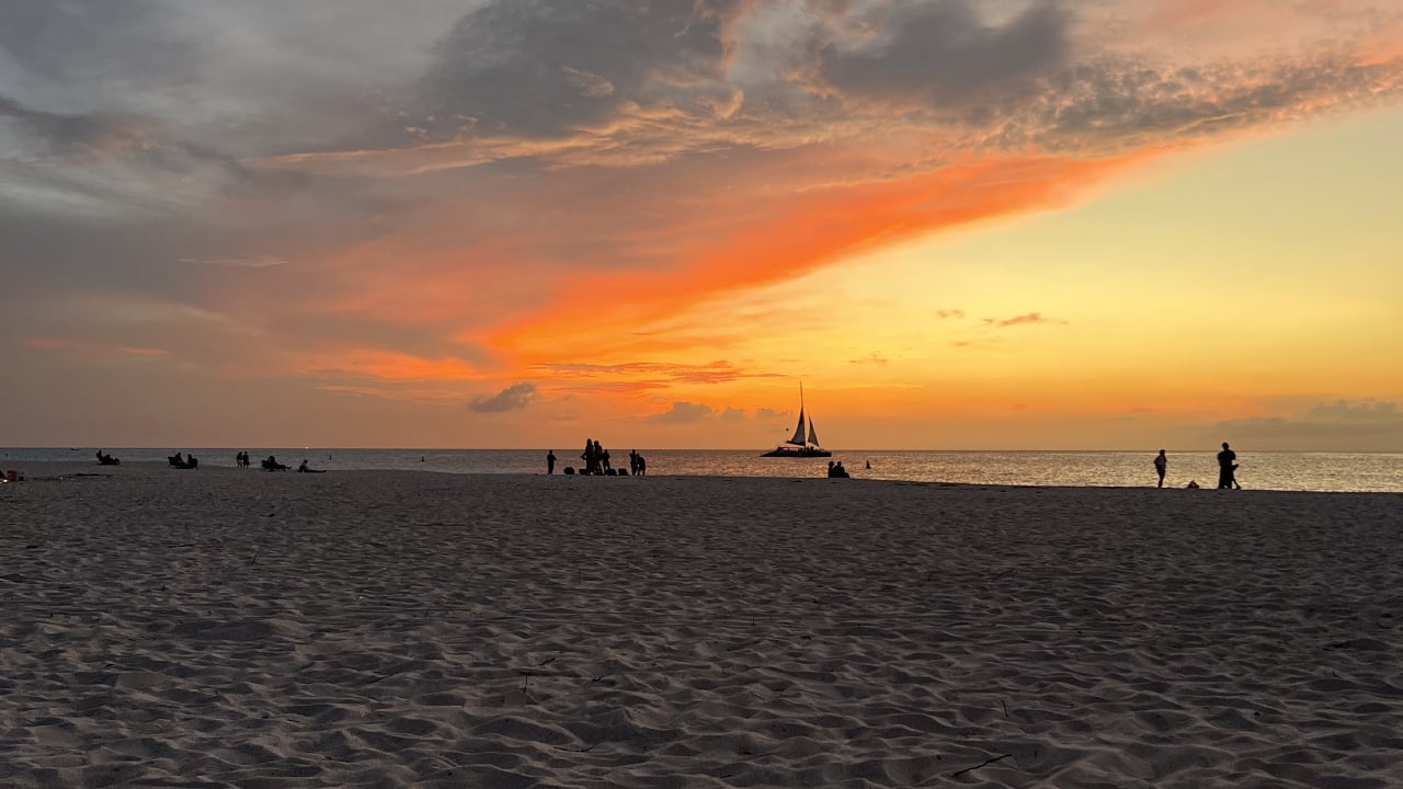 Un atardecer en Eagle Beach tiñe el cielo de tonos naranjas, amarillos y grises, mientras el sol se esconde sobre el mar sereno y las siluetas de los veleros y la gente contrastan