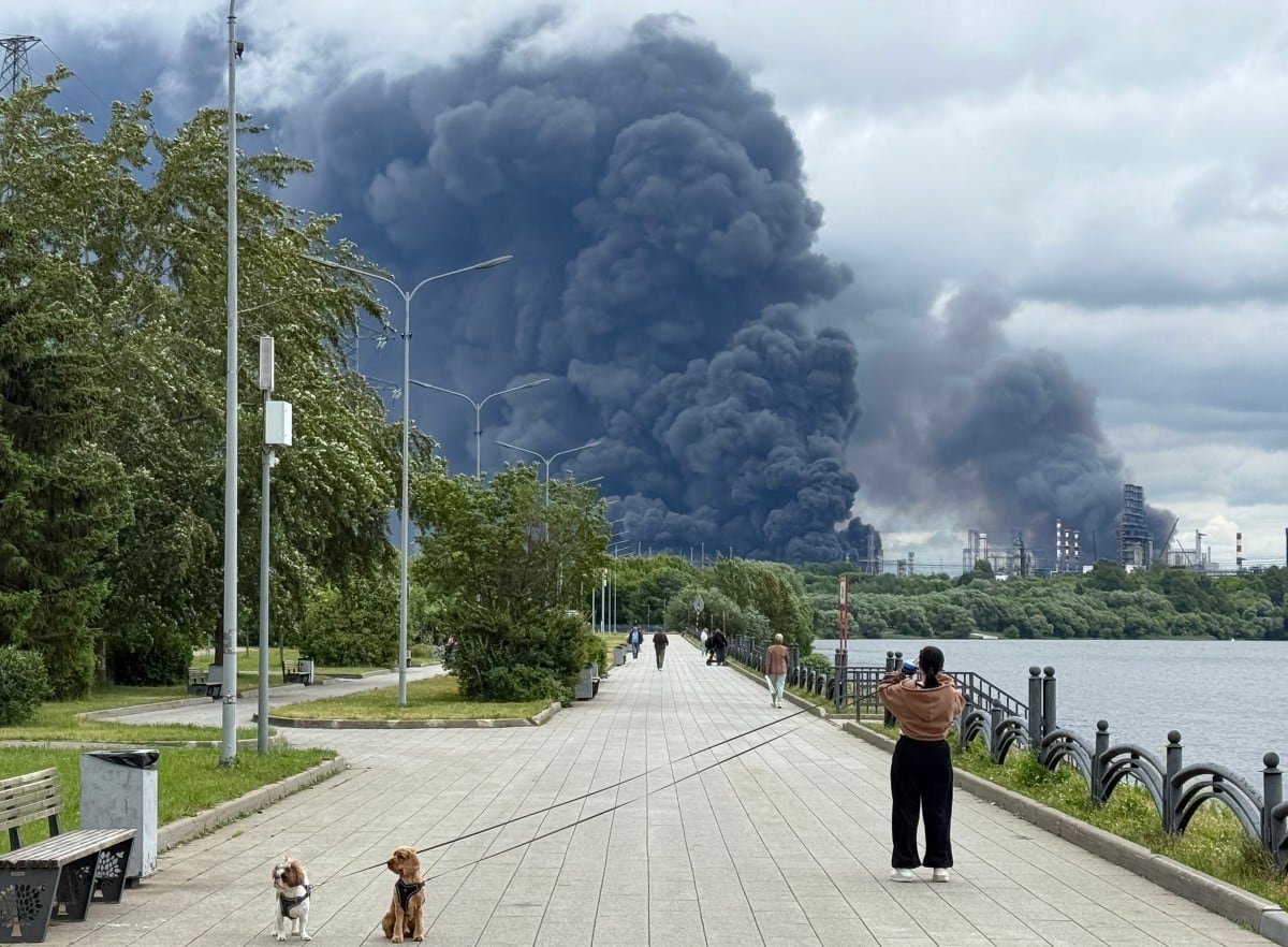 Varias personas pasean por un parque mientras una densa columna de humo negro se eleva desde la refinería de petróleo de Gazprom Neft, la petrolera rusa ubicada en las afueras del sureste de Moscú, el 18 de junio de 2026.