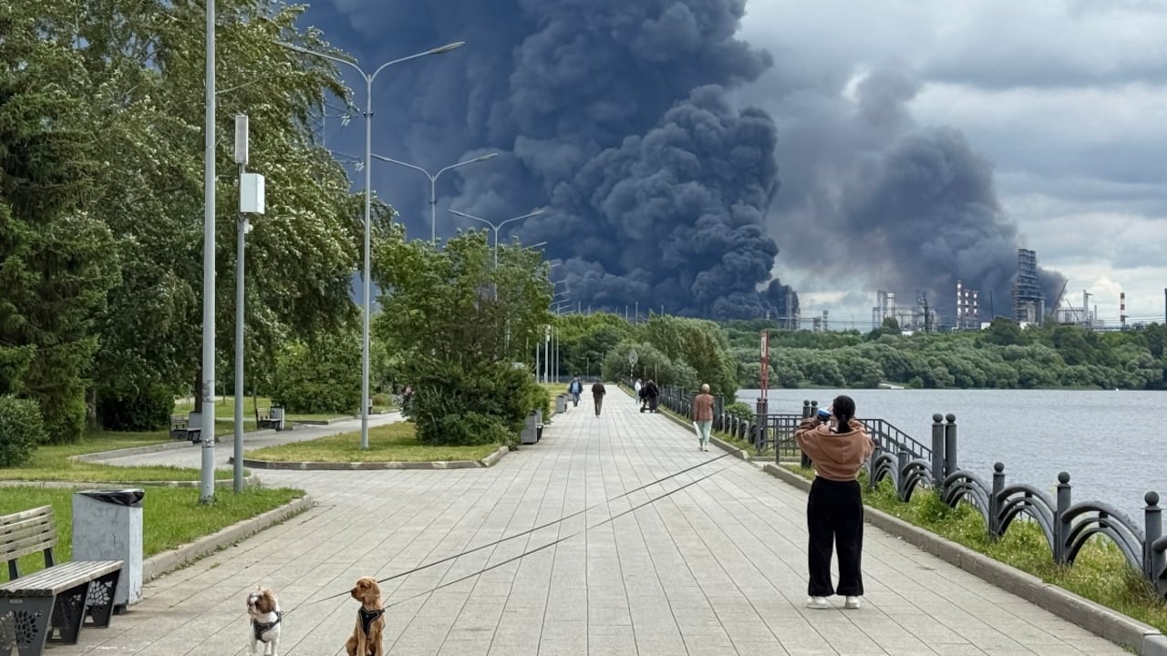 Varias personas pasean por un parque mientras una densa columna de humo negro se eleva desde la refinería de petróleo de Gazprom Neft, la petrolera rusa ubicada en las afueras del sureste de Moscú, el 18 de junio de 2026.