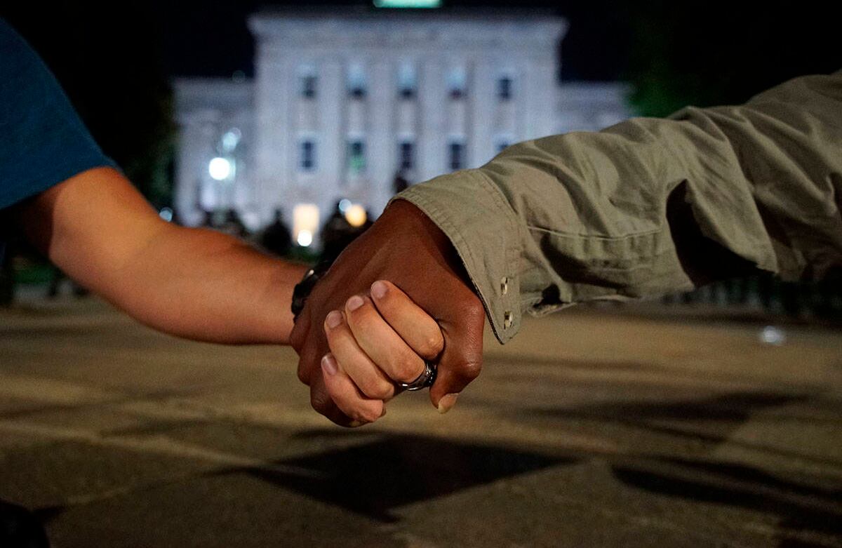 Un hombre negro y uno blanco se dan la mano mientras la policía en el fondo vigila el antiguo capitolio estatal en Raleigh, Carolina del Norte, el lunes 1 de junio de 2020. Era el segundo día de protestas en la capital de Carolina del Norte tras la muerte de George Floyd mientras estaba bajo custodia policial. Foto: Allen G. Breed / AP.