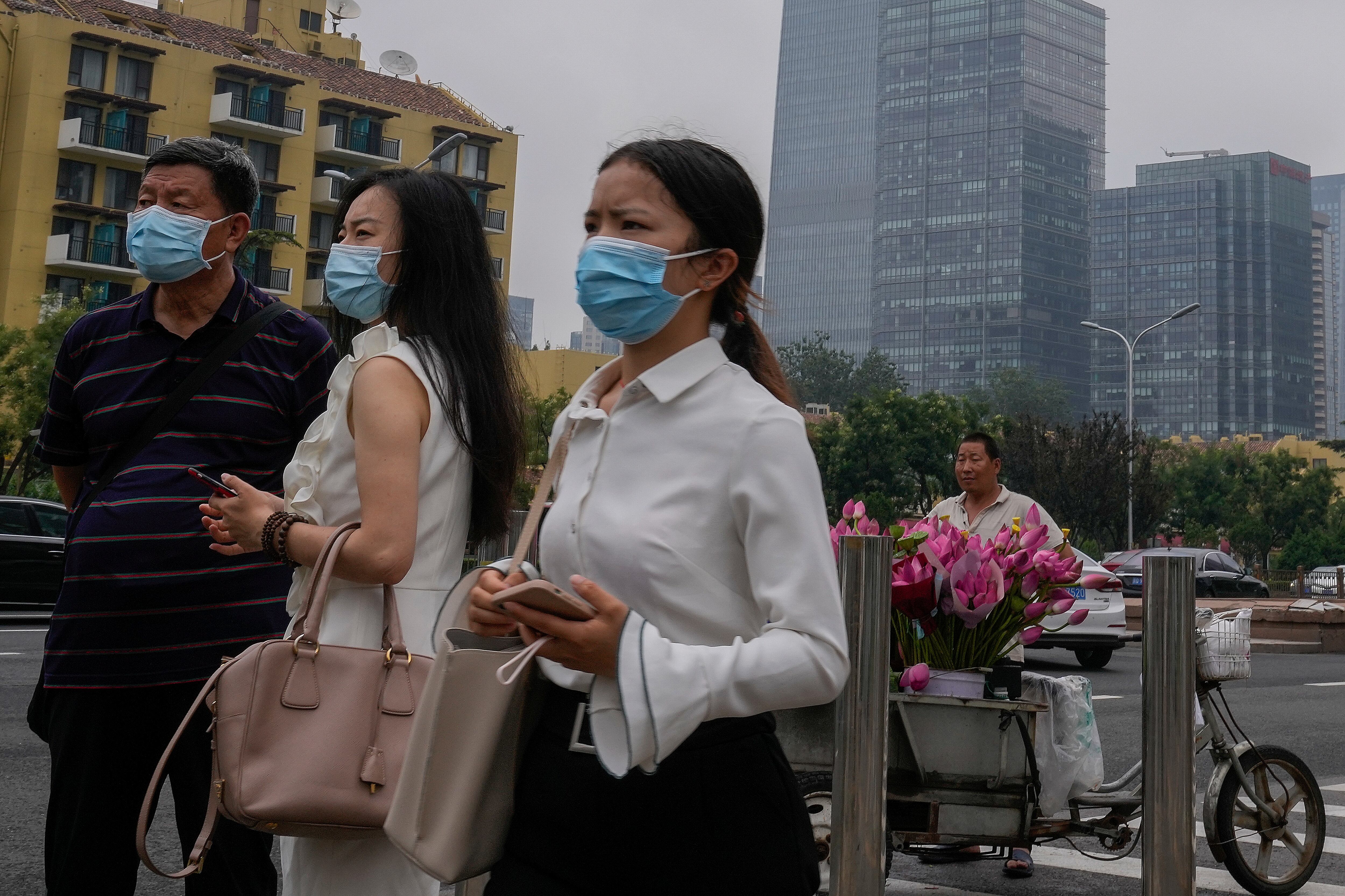 Varias personas con mascarillas para protegerse del coronavirus observan algo junto a un vendedor de flores afuera de una estación del tren subterráneo en Beijing. (AP Foto/Andy Wong)