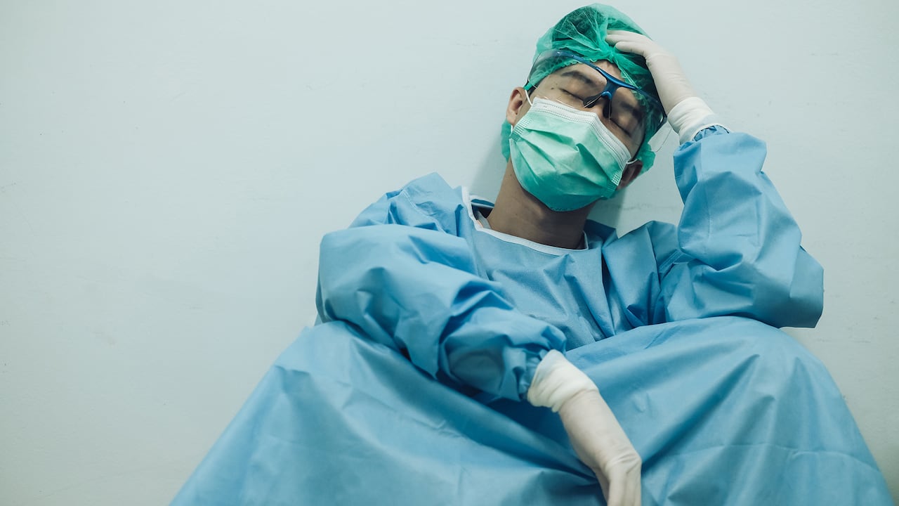 Médico con dolor de cabeza y cansado del trabajo mientras usa traje de PPE para proteger la enfermedad del coronavirus. El bienestar y la resiliencia emocional son componentes clave para mantener los servicios de atención esenciales.