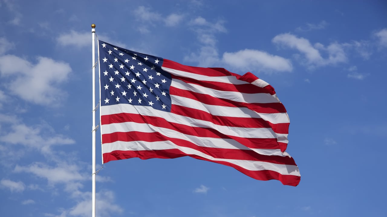 Bandera estadounidense ondeando al viento, con cielo azul y nubes detrás.