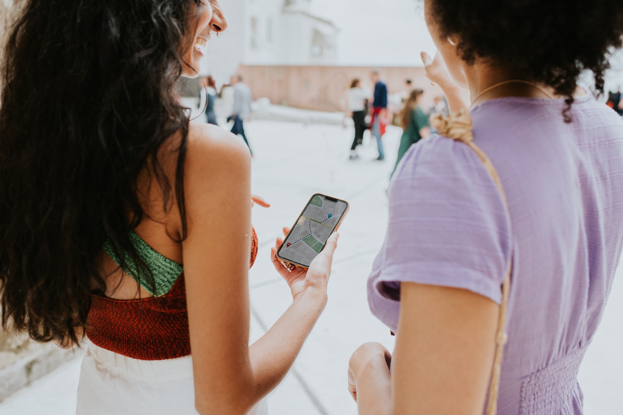 A woman on vacation uses her smart phone for directions on foot. A female friend stands beside her and discusses the route. Tourists are visible in the background. Depicts roaming, phone plans, travel and map applications. Space for copy.