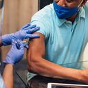 Una enfermera de raza mixta con un protector facial, mascarilla quirúrgica y guantes protectores que administra la vacuna COVID-19 a un hombre negro de alto nivel en su casa.