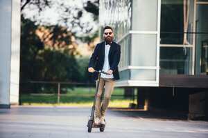 Un hombre guapo, joven y barbudo con un traje montado en un scooter eléctrico frente a un edificio de oficinas.