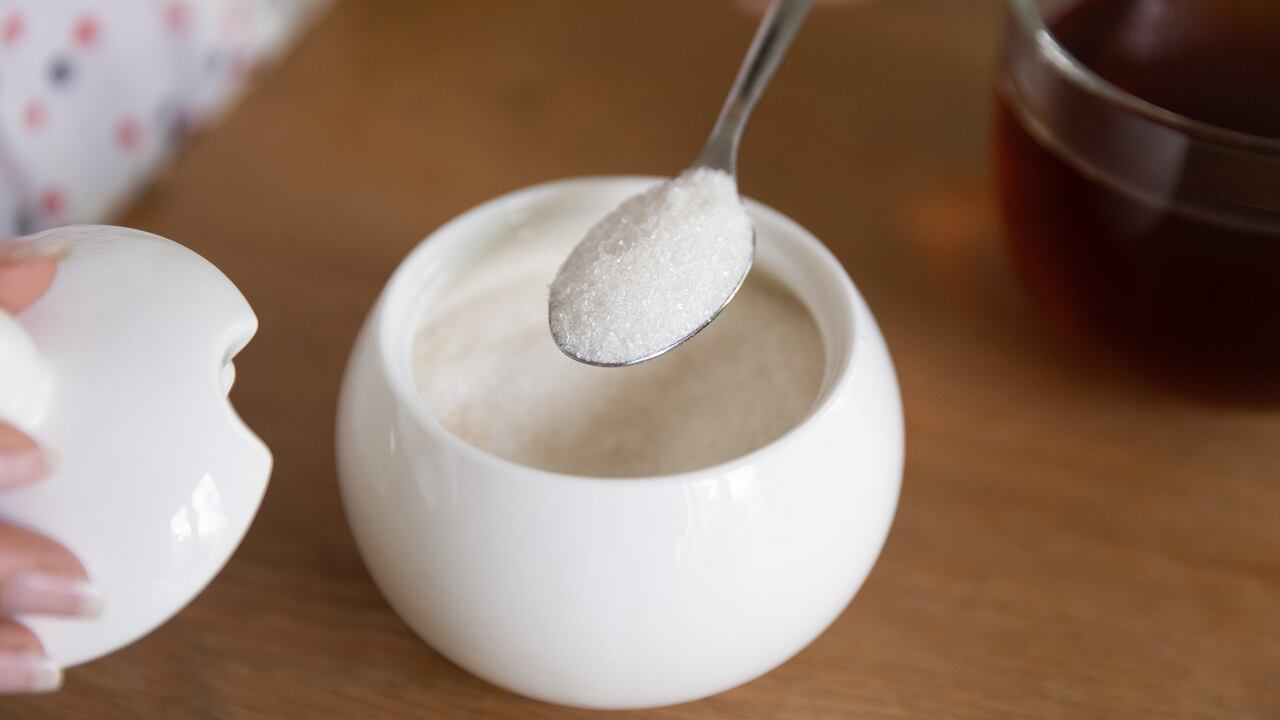 Primer plano de una mujer poniendo una cuchara de azúcar granulada en té negro de un tazón de porcelana blanca, una joven disfrutando de la rutina matutina diaria, preparando el desayuno en casa, bebiendo bebidas calientes