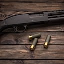 A black 12-gauge pump-action shotgun and ammo are on a wooden table. A smooth-bore weapon with a plastic stock. Dark brown background.