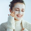 smiling stylish woman in roll neck sweater and cardigan applying white facial mask isolated on winter light blue background.