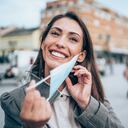 Hermosa joven feliz con máscara facial médica protectora para la protección contra virus durante la pandemia de Coronavirus/COVID-19. Mujer joven poniendo/quitando su máscara al aire libre en la ciudad.