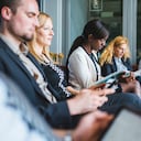 Grupo de empresarios sentados y esperando una entrevista de trabajo. Están utilizando teléfonos móviles y tabletas digitales. Foto: Getty Images.