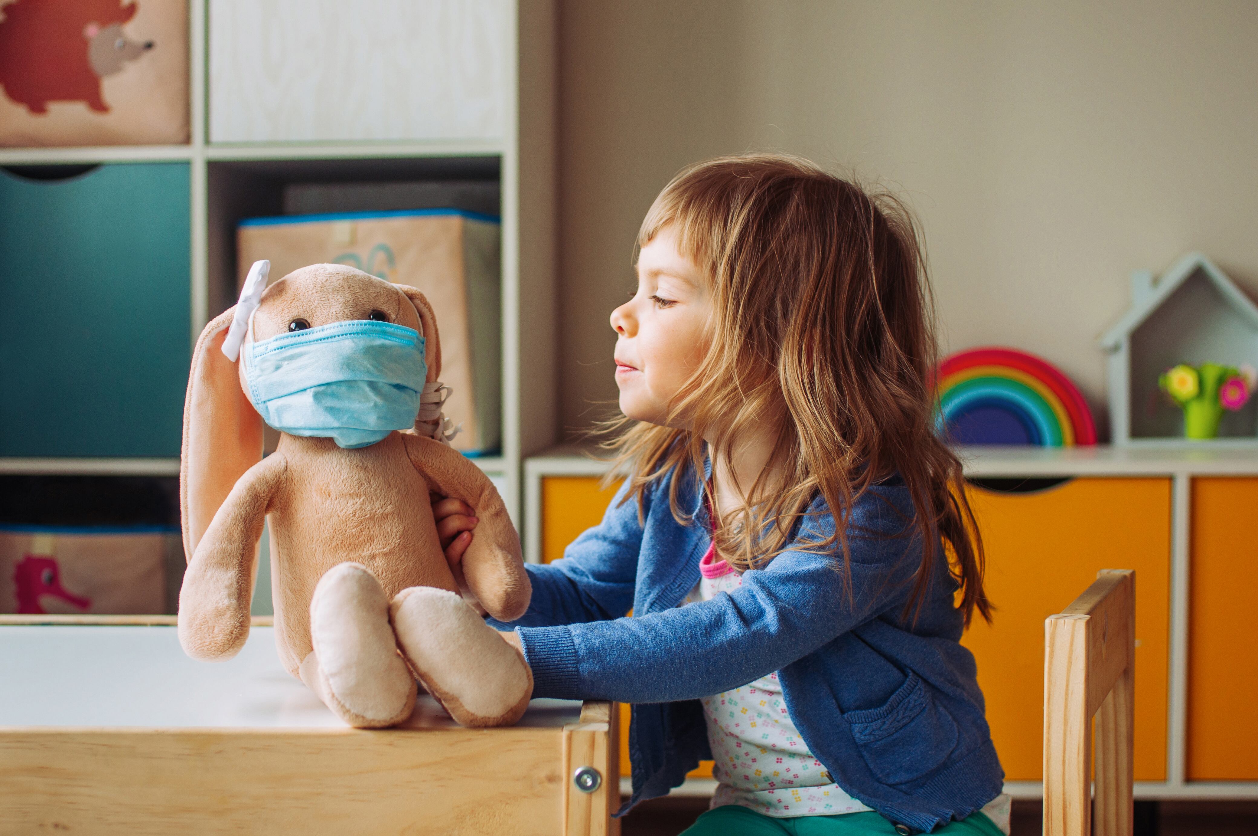 Niña jugando con peluche de conejo en la máscara de la medicina sentado en la mesa en la habitación de los niños.