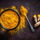 Spices: Top view of a black bowl filled with turmeric powder shot on abstract brown rustic table. A metal spoon with turmeric powder is beside the bowl and turmeric powder is scattered on the table. Fresh organic turmeric roots are beside the spoon. Predominant colors are brown and yellow. Low key DSRL studio photo taken with Canon EOS 5D Mk II and Canon EF 100mm f/2.8L Macro IS USM.