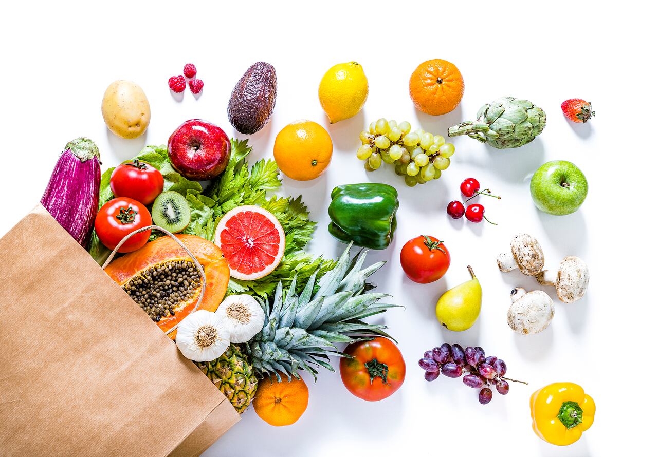 Top view of a paper bag full of various kinds of multicolored fruits and vegetables like papaya, tomatoes, red and green apples, carrots, mushrooms, eggplants, pineapple, cherries, lime, garlic, oranges and kiwi. The paper bag is laying at the lower left corner on a white background and the fruits and vegetables are coming out from it. Studio shot taken with Canon EOS 6D Mark II and Canon EF 24-105 mm f/4L.