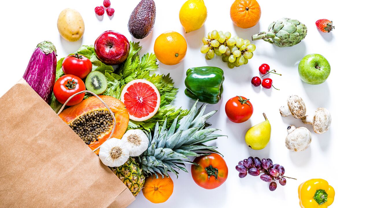 Top view of a paper bag full of various kinds of multicolored fruits and vegetables like papaya, tomatoes, red and green apples, carrots, mushrooms, eggplants, pineapple, cherries, lime, garlic, oranges and kiwi. The paper bag is laying at the lower left corner on a white background and the fruits and vegetables are coming out from it. Studio shot taken with Canon EOS 6D Mark II and Canon EF 24-105 mm f/4L.