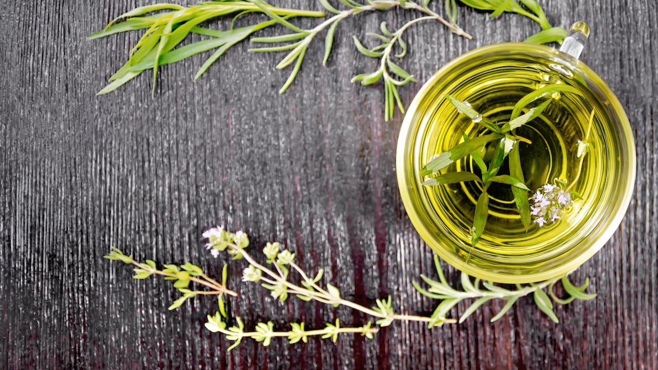 Frame with herbal tea in a glass cup, sprigs of tarragon, rosemary and thyme on wooden board background from above