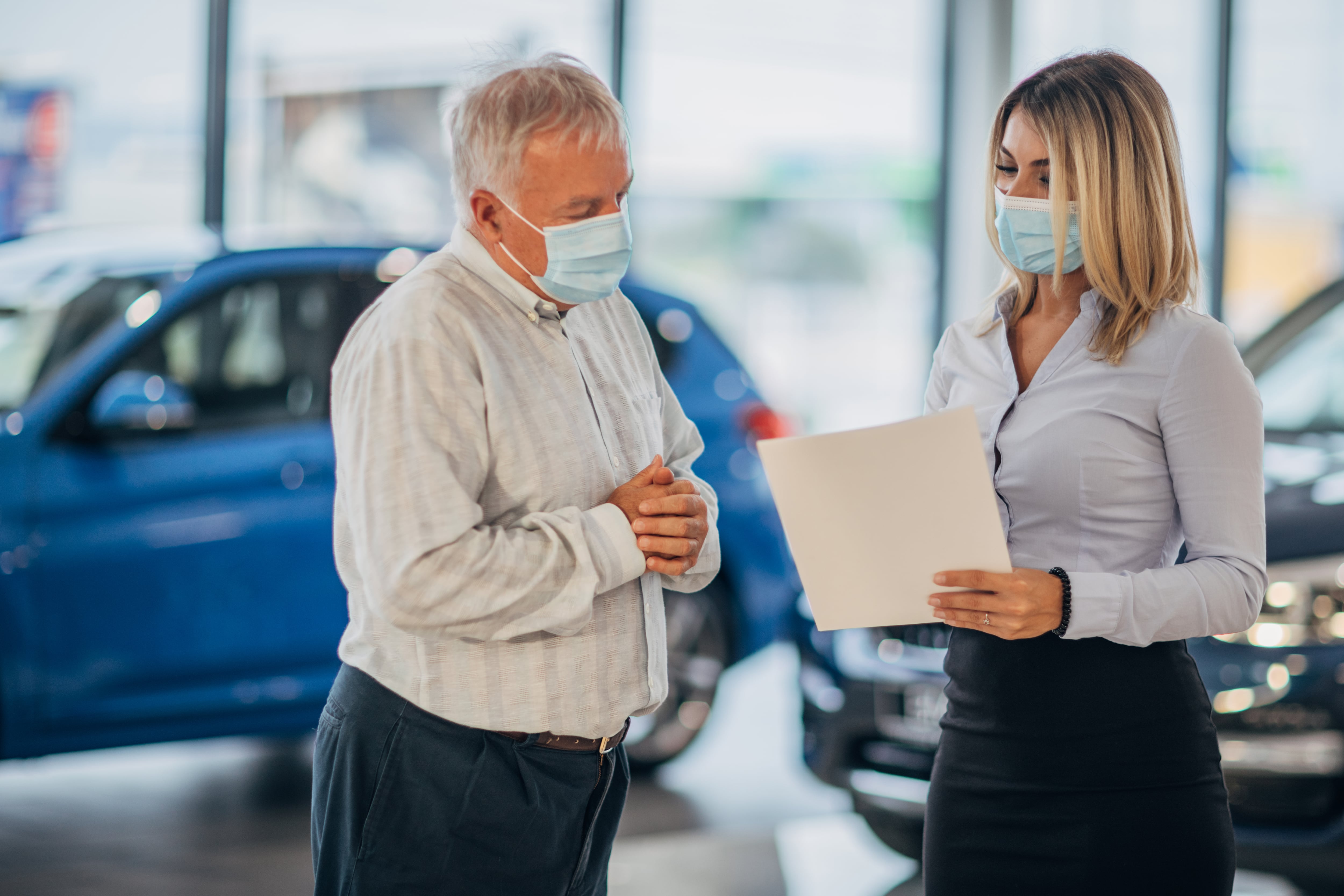Female car dealer and senior man with protective face masks going through paperwork in a showroom