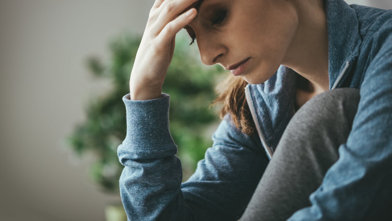 Sad depressed woman at home sitting on the couch, looking down and touching her forehead, loneliness and pain concept