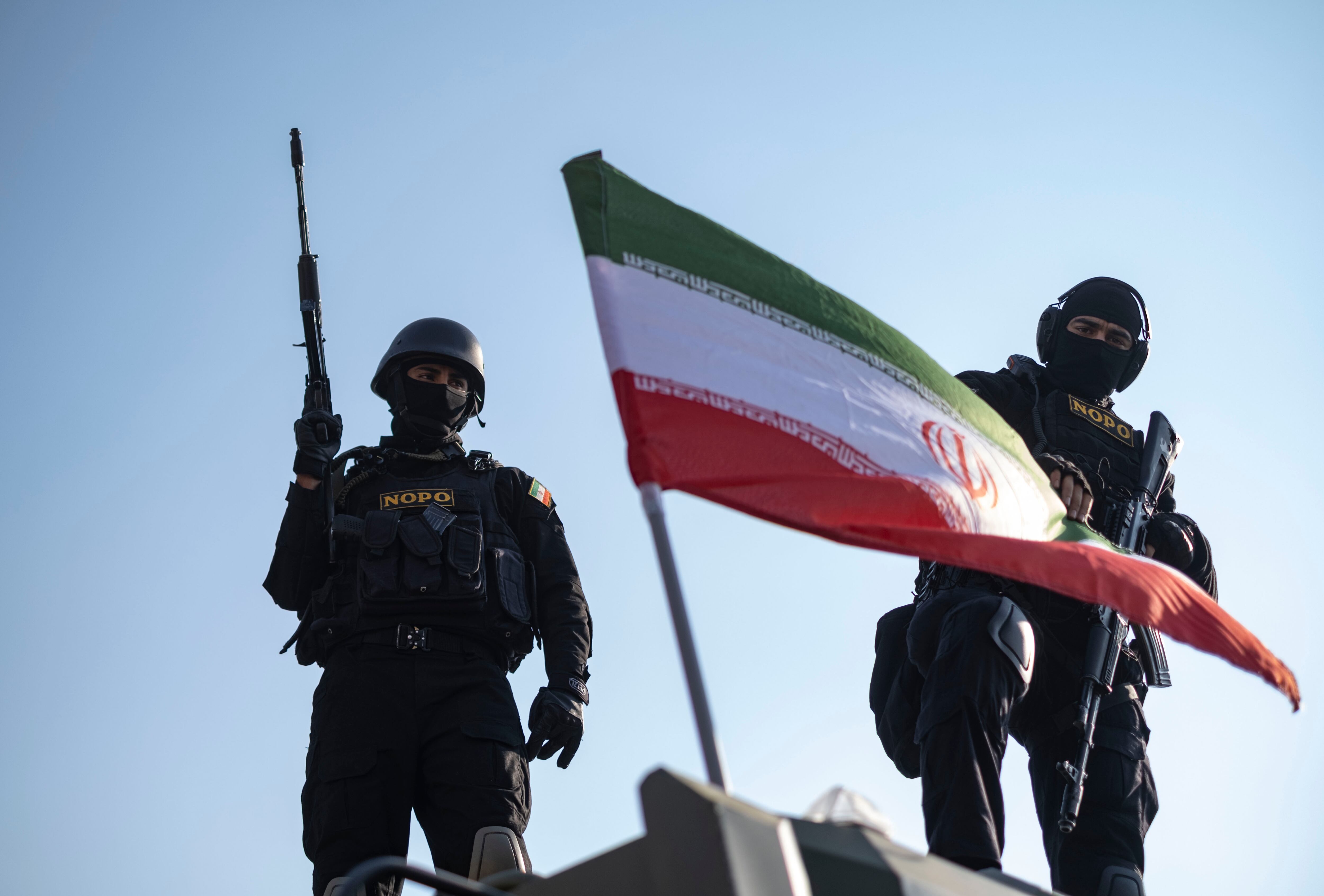 Dos miembros armados de las fuerzas especiales de la policía iraní posan detrás de una bandera iraní colocada en un vehículo militar blindado