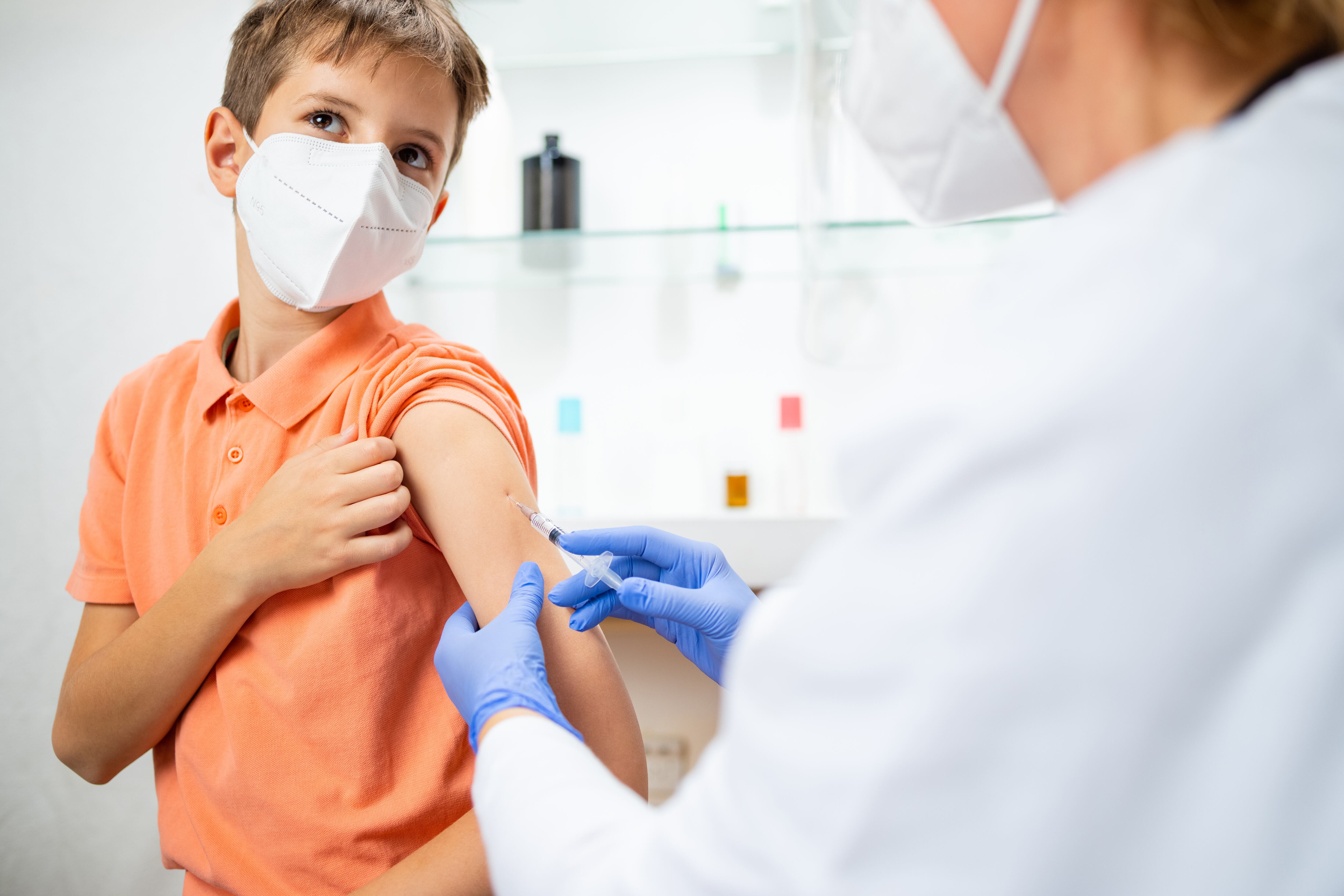 Niño con mascarilla protectora sosteniendo la manga de su camisa y mirando hacia otro lado para no mirar al médico inyectando la vacuna en su brazo