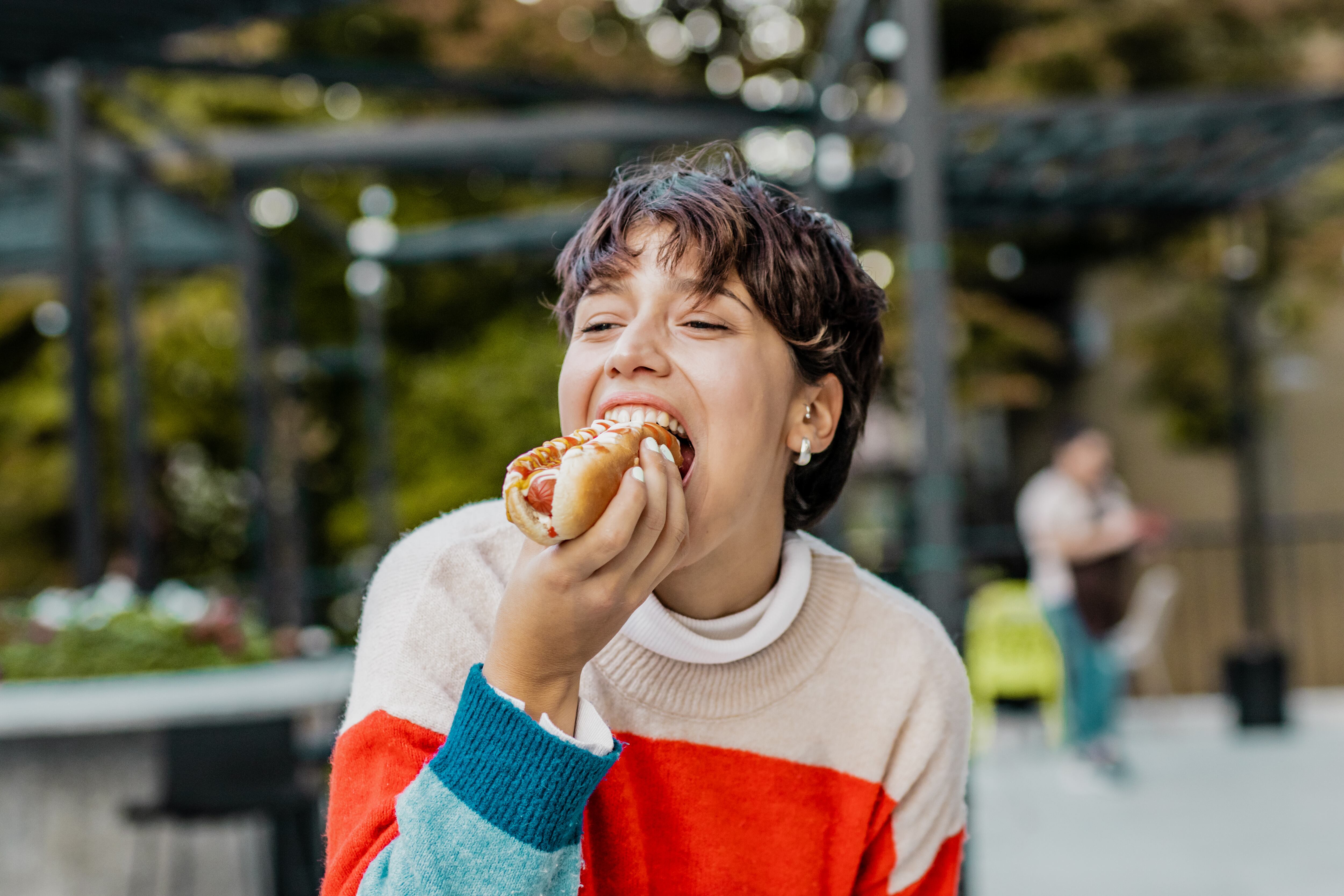 Mujer joven comiendo varita de hot dog riendo en la calle