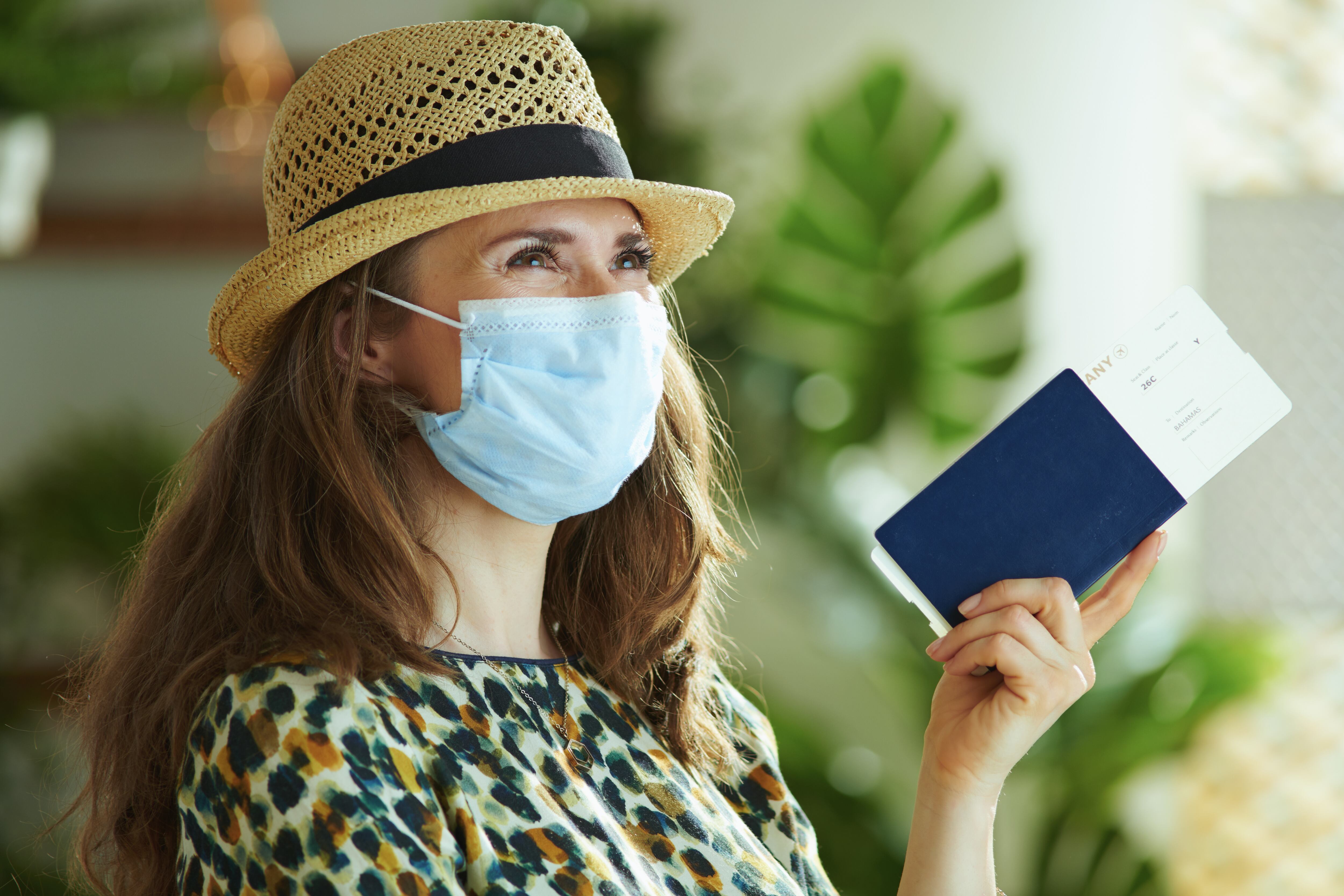 La vida durante la pandemia del covid-19. mujer de moda soñadora con sombrero de paja con máscara médica, pasaporte y boleto aéreo mirando en la distancia.