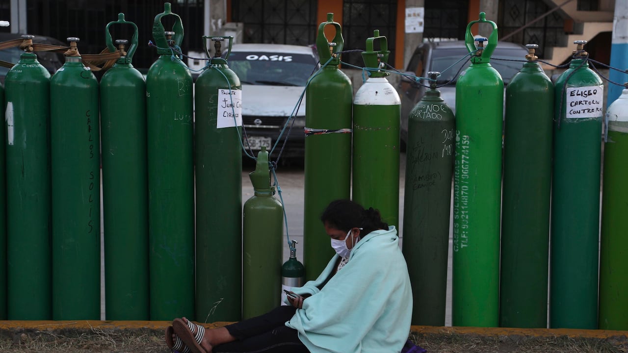 Una mujer revisa su teléfono celular mientras se sienta junto a su tanque de oxígeno vacío después de acampar durante la noche para esperar con otros a que abra una tienda de recargas en Callao, Perú. (AP Foto/Martín Mejía)
