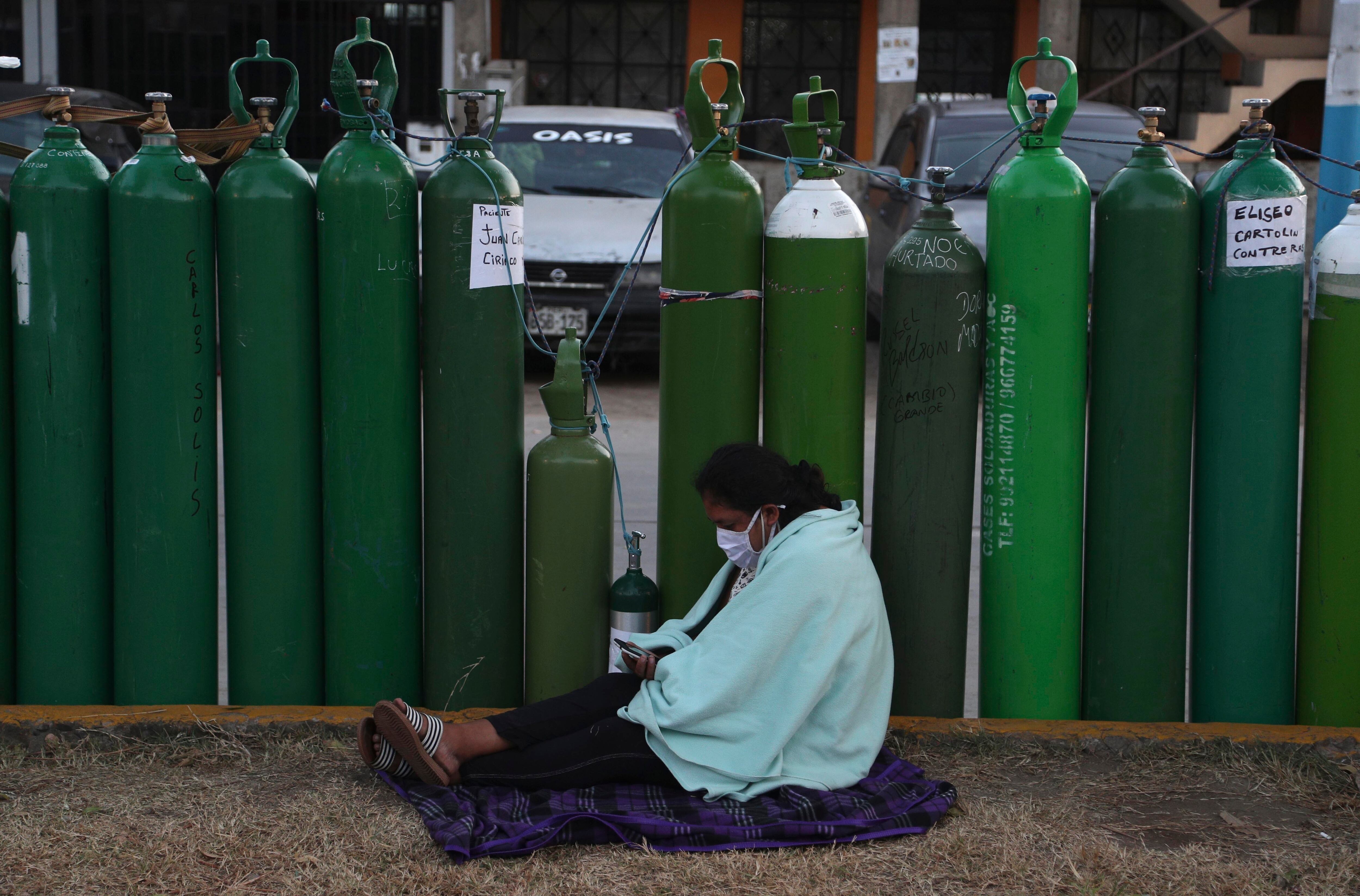 Una mujer revisa su teléfono celular mientras se sienta junto a su tanque de oxígeno vacío después de acampar durante la noche para esperar con otros a que abra una tienda de recargas en Callao, Perú. (AP Foto/Martín Mejía)