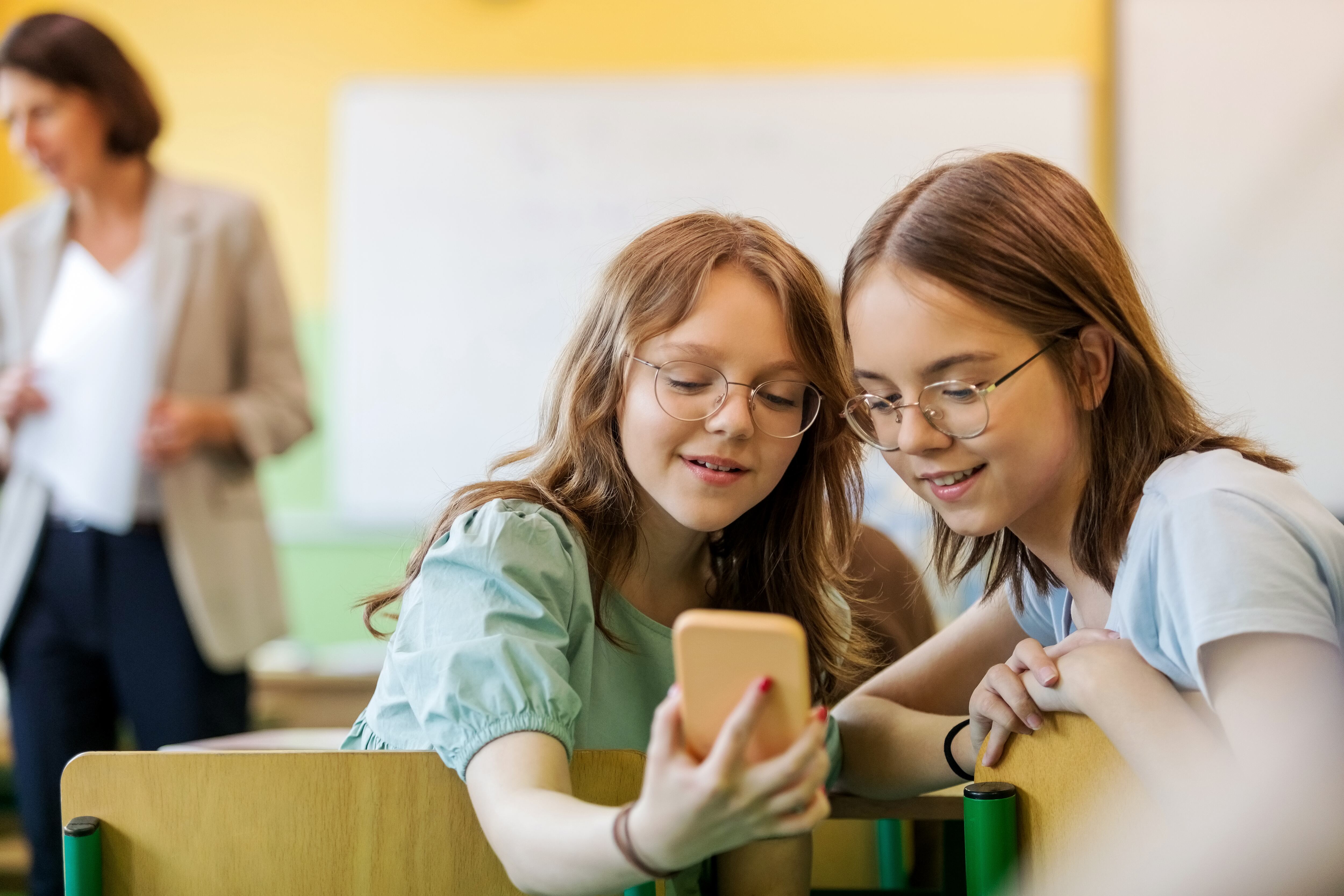 Estudiantes de secundaria sentadas en el escritorio del aula y usando teléfonos inteligentes con un profesor desenfocado en el fondo.