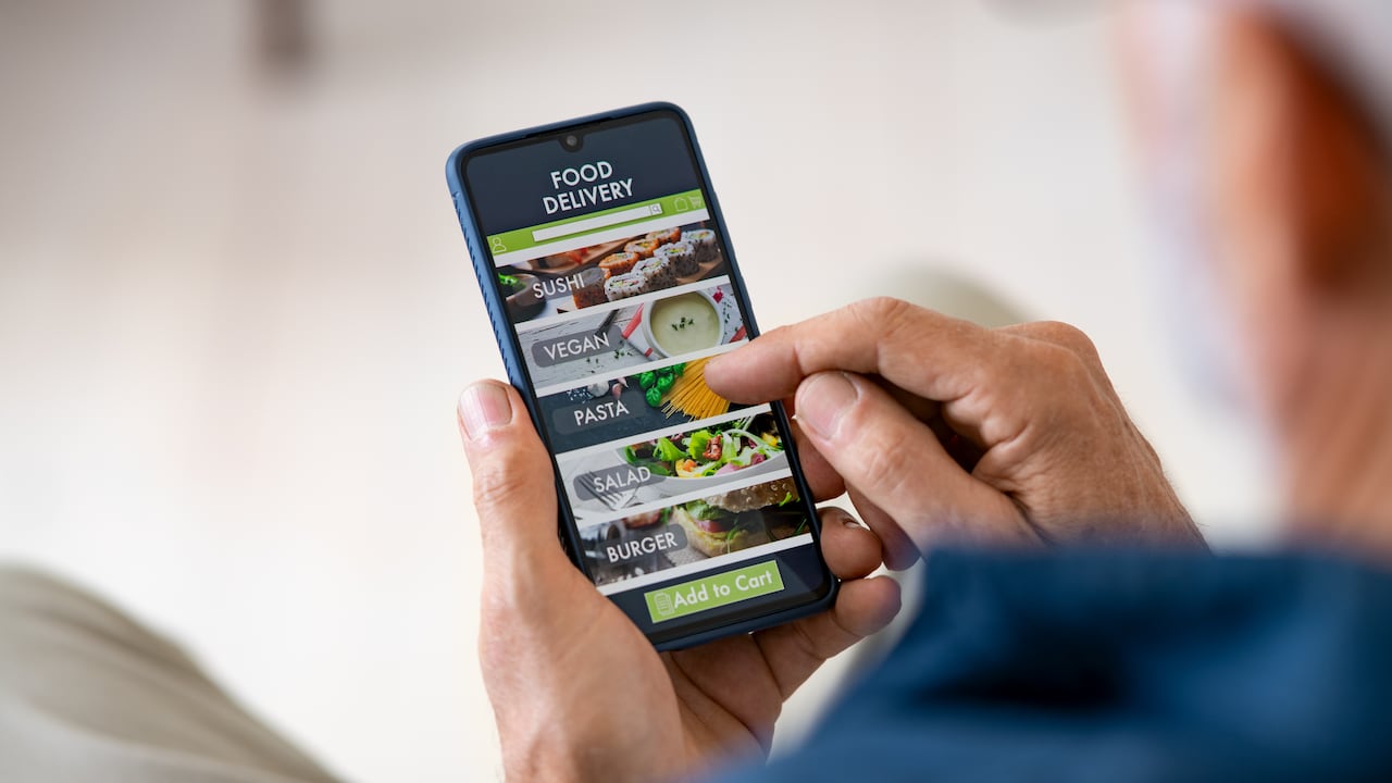 Vista superior de las manos del hombre sosteniendo el teléfono inteligente mientras pide la entrega de alimentos en casa. Vista posterior del hombre maduro que usa la aplicación de entrega de alimentos con el teléfono celular para pedir el almuerzo.