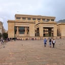Mirando hacia la esquina noreste de la Plaza Bolívar en la capital andina de Bogotá en Colombia, Sur América. El edificio a la vista es el 'Palacio de Justicia' o traducido, el Palacio de Justicia. Es la Corte Suprema del País. Muchos colombianos locales pasan despreocupadamente, mientras realizan su día a día mientras conversan con amigos o familiares. Como muchas otras plazas en todo el mundo, hay muchas palomas en la plaza esperando a que los visitantes las alimenten. A la derecha de la imagen se puede ver el pico andino de Monserrate. El cielo está nublado; lloverá en breve. La altitud al nivel de la calle es de unos 8.500 pies sobre el nivel del mar. La mayoría de las personas usan una chaqueta o una capa adicional de ropa para mantenerse abrigados. Foto tomada a la luz del sol de la tarde en un día nublado; formato horizontal.