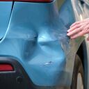 Driver hand examining dented car with damaged fender parked on city street side. Road safety and vehicle insurance concept.