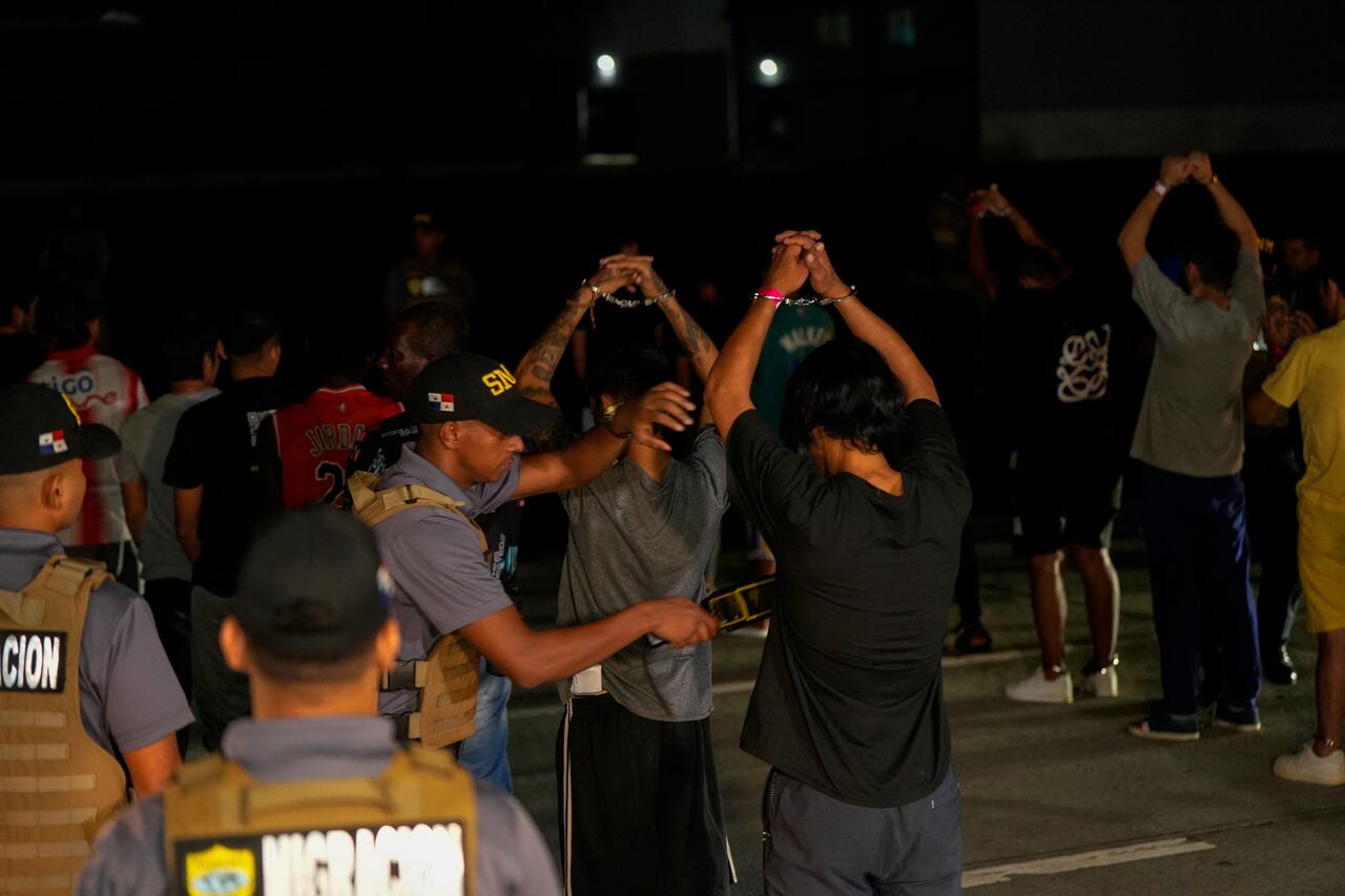 Migrantes colombianos esposados se alinean al costado de la pista del aeropuerto de Albrook Gelabert en la ciudad de Panamá el 20 de agosto de 2024, mientras son examinados con detectores de metales antes de su deportación. Panamá deportó este martes a 29 colombianos con antecedentes penales que habían ingresado al país a través del inhóspito Tapón del Darién, aplicando por primera vez un acuerdo sobre repatriaciones de migrantes firmado con Estados Unidos en julio. (Foto de ARNULFO FRANCO / AFP)