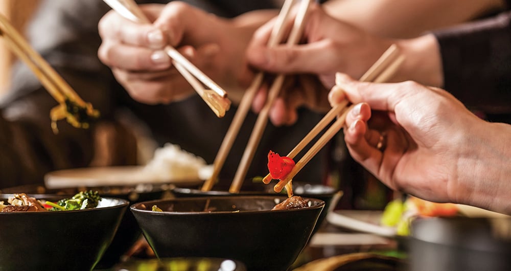 Four sets of chopsticks grabbing Asian food out of various black bowls sitting on a table in a Thai food restaurant.  Three pairs of chopsticks have food between them, and one is in a black bowl.  There is a person's chest out of focus in the background.  There are four sets of chopsticks visible, but there are only three hands visible.  There is a set of chopsticks coming into view from the left.  The hand in front is in focus, while the two hands in the background are slightly blurred.