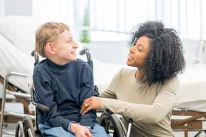 Un niño pequeño está sentado en su silla de ruedas con su cuidador. Están sentados juntos en el hospital y están sonriendo.