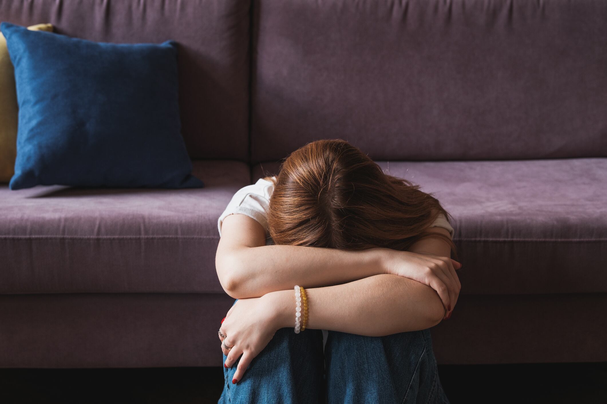 Upset depressed teenager covering face with hands and crying, sad frustrated teen girl sitting on floor near sofa at home, having problems. Teenagers and mental health concept