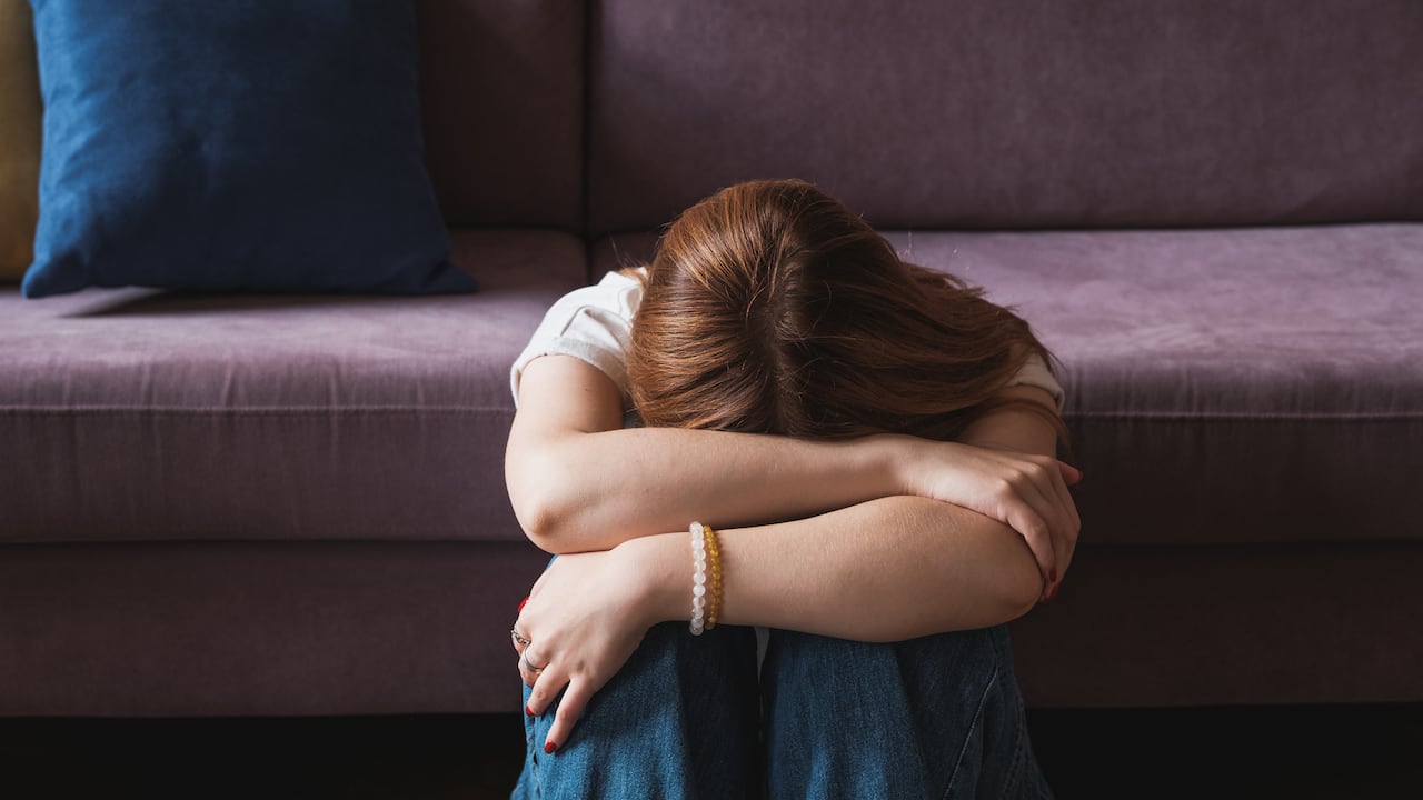 Upset depressed teenager covering face with hands and crying, sad frustrated teen girl sitting on floor near sofa at home, having problems. Teenagers and mental health concept