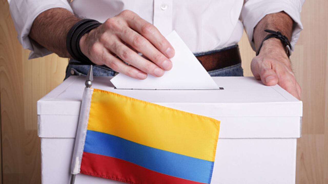 A man inserting a ballot to a ballot box. Colombian flag in front of it.