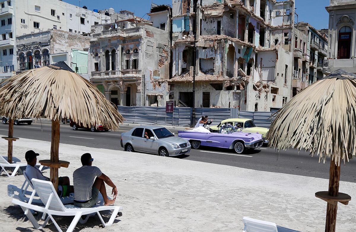 Las personas se sientan en las sillas de una playa artificial, que hace parte de la intervención artística 'Detrás del muro', mientras unos recién casados pasan en un carro convertible en el Malecón habanero, Cuba. (AP/Desmond Boylan)