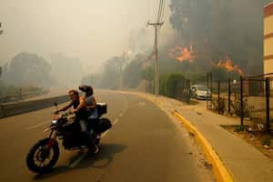 Residents evacuate on a motorcycle as smoke caused by forest fires fill the sky and flames spread into Vina del Mar, Chile, Saturday, Feb. 3, 2024. Officials say intense forest fires burning around a densely populated area of central Chile have left several people dead and destroyed hundreds of homes. (AP Photo/Esteban Felix)