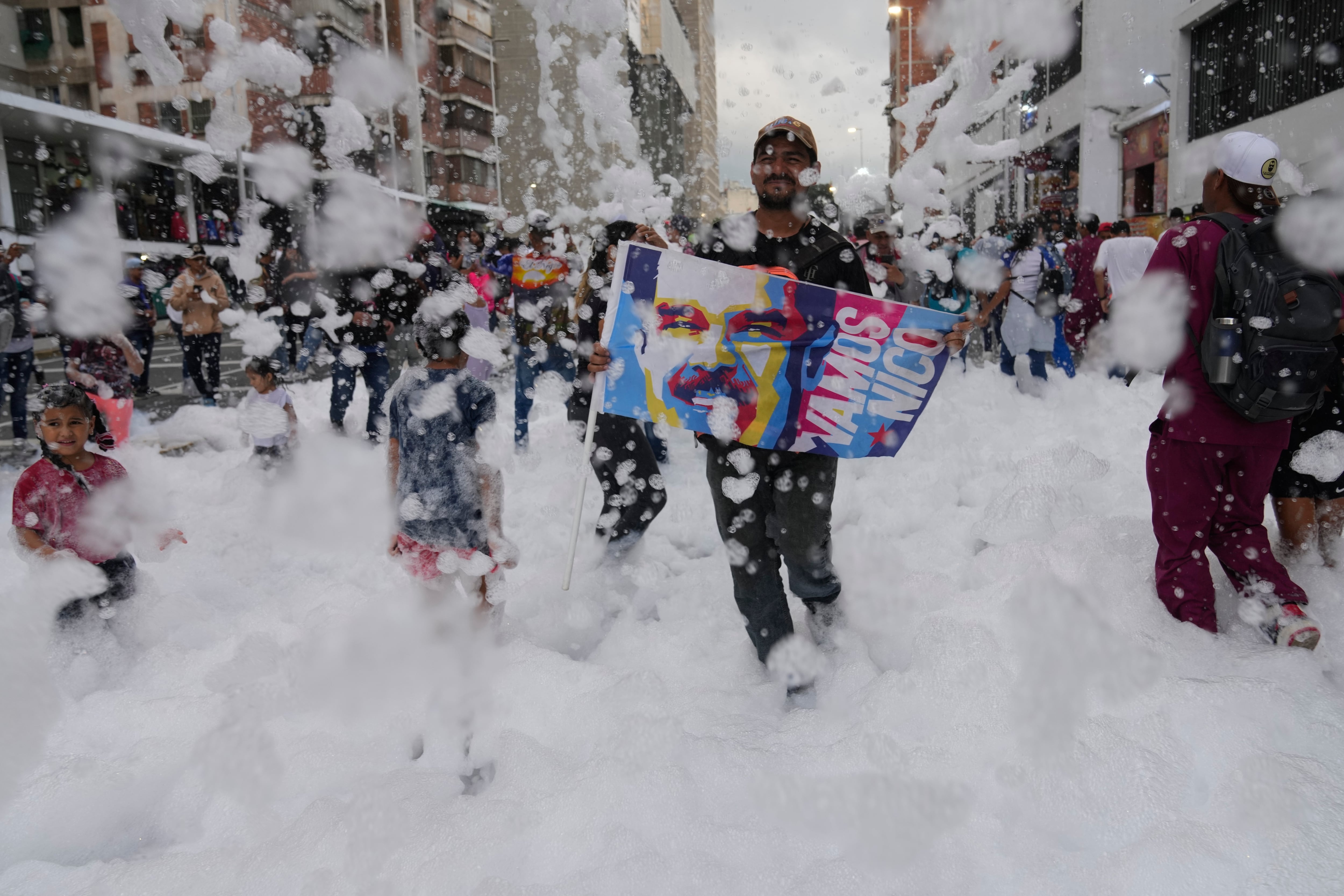 A person holding a poster of Venezuelan President Nicolas Maduro walks through foam sprayed for an event celebrating University Student Day in Caracas, Venezuela, Thursday, Nov. 21, 2024. (AP Photo/Ariana Cubillos)