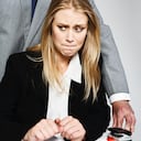 Un hombre de negocios se para detrás de una mujer de negocios joven y bonita, rodeándola con los brazos mientras se sienta en su escritorio con aspecto amenazado, aterrorizado e indefenso.