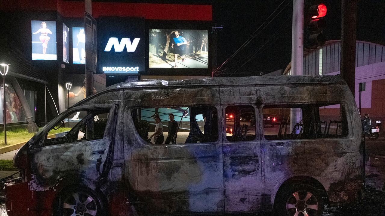 Personas observan un vehículo de transporte quemado tras ser incendiado por individuos no identificados en Tijuana, estado de Baja California, México, el 12 de agosto de 2022. Varios vehículos fueron incendiados, ese mismo día, en la ciudad. (Photo by Guillermo Arias / AFP)